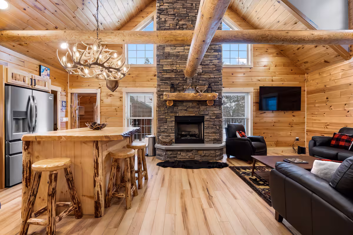 Great room in the cabin with vaulted pine ceiling, antler chandelier, live-edge kitchen island, and a full stone fireplace