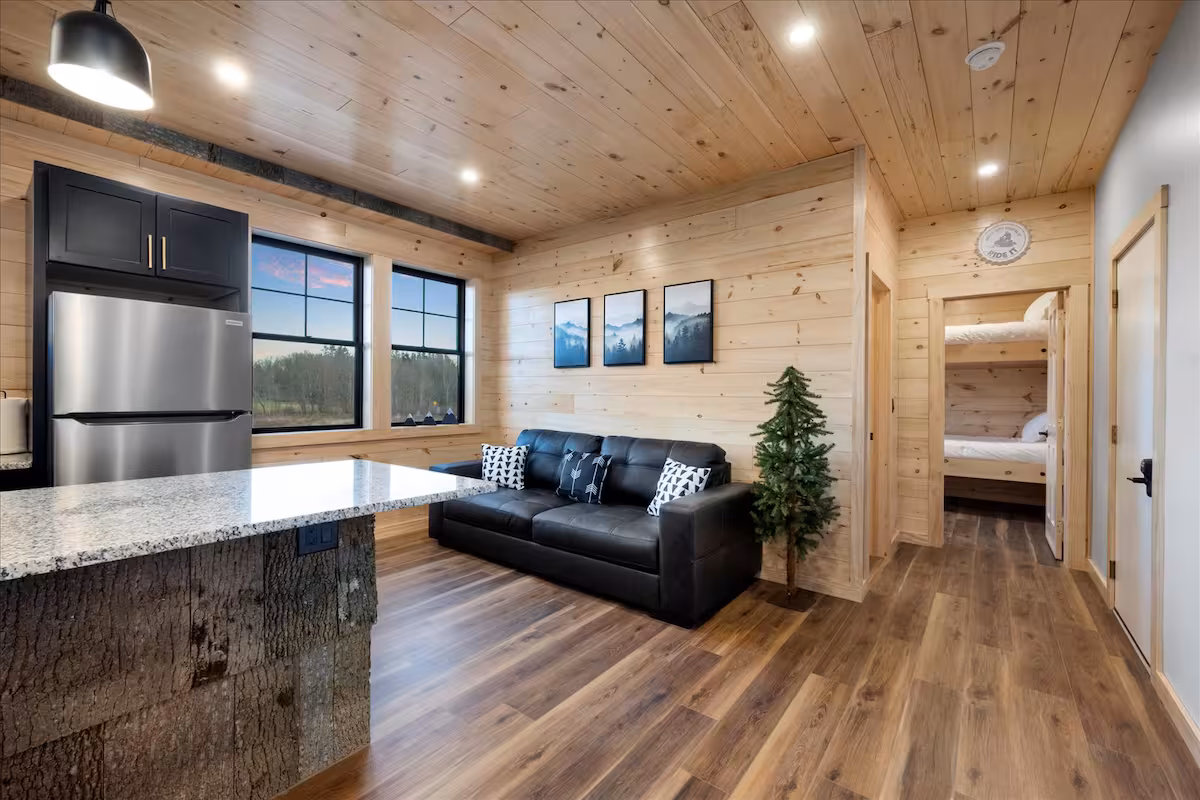 Open kitchen and living area with black cabinetry, a stone-clad island, and a black leather sofa, bunkroom visible through the doorway