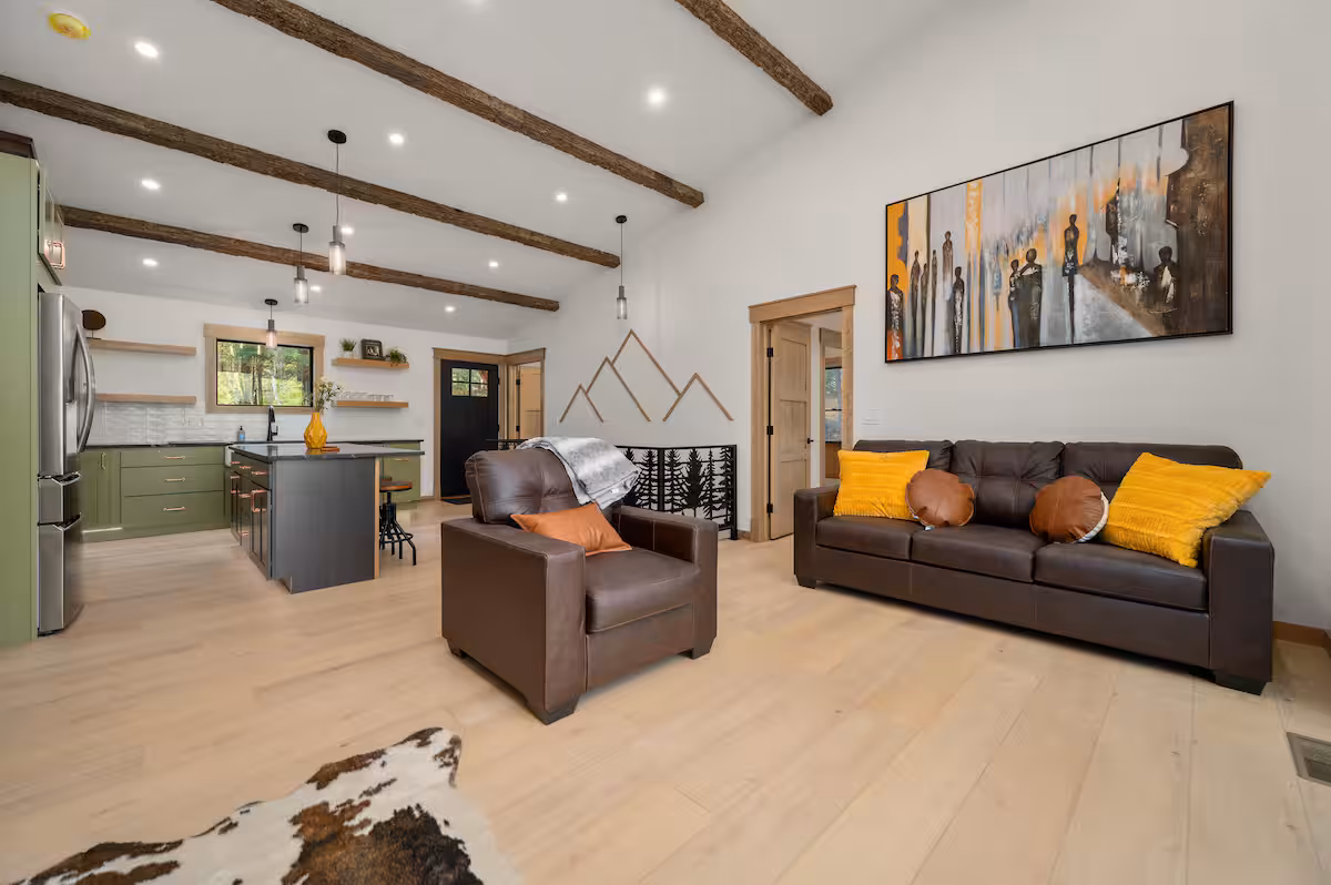 Vaulted great room with olive-green kitchen cabinetry, exposed wood beams, a leather sofa and armchair, and a cowhide rug