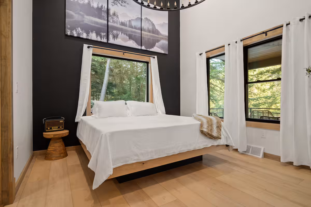 Primary bedroom with a floating platform bed, round chandelier, and tall windows looking out over the trees and porch